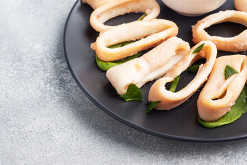 Fried squid rings with tomato sauce and lemon. Black plate, grey concrete background copy space.