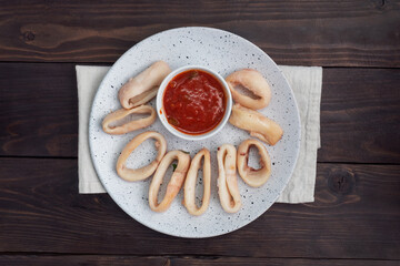 Fried squid rings with tomato sauce and lemon. Dark wooden background copy space. Top view.