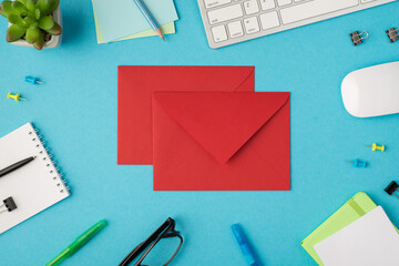 Top view photo of workplace with chancellery keyboard mouse glasses plant and closed red envelopes in the middle on isolated blue background