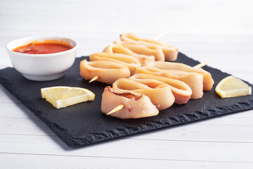 Fried squid rings on a skewer with tomato sauce and lemon. Black slate stand, white background copy space.