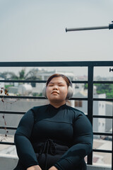 Peaceful plus size young asian woman in modern wireless headphones sit relax after doing exercise on mat in balcony, going to loose extra pounds at home, become strong and fit.