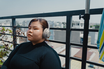 Peaceful plus size young asian woman in modern wireless headphones sit relax after doing exercise on mat in balcony, going to loose extra pounds at home, become strong and fit.