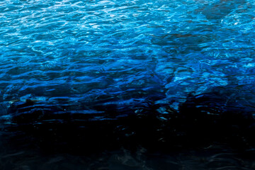 Fototapeta na wymiar Water in a pool with slightly moving waves and reflections of the sun, gradient from light to dark blue to black with copy-space