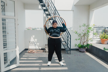 Plus size young asian woman playing hula hoop in balcony. Helps to burn excess fat in body. Strengthens the muscles of the body, going to loose extra pounds at home,