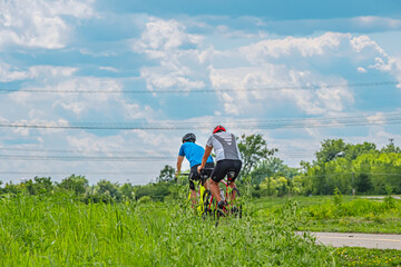 Two high energy colorfully dressed cyclists prepare to pass under high energy wires while on a nature cycle path. The cycle path is framed by green fields and a blue cloud filled sky