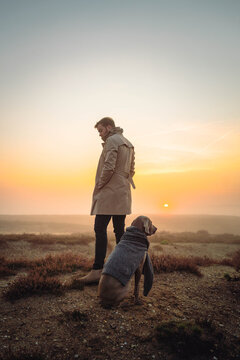A young man in a beige coat standing in the foggy field with a dog by side at the sunrise