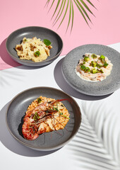 Seafood risotto with shrimp. Mushroom risotto and creamy with avocado. RIsotto plates on white table and pink table. Sunlight with harsh shadow of green tropic leaf