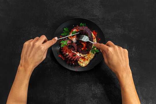 Anonymous person cutting a Grilled Octopus with hummus and spices on a dark background