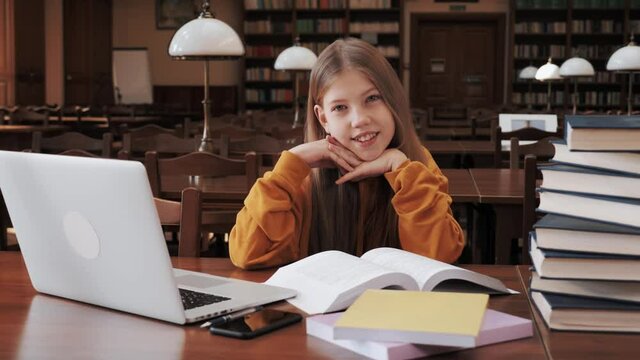 Beautiful teenage schoolgirl in a yellow sweater sitting at a table in a classic library and doing homework, she reads a book and uses a laptop with the Internet to access the book database education.