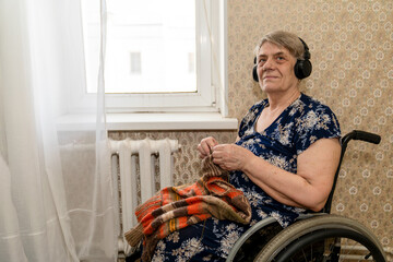 an elderly woman with headphones sits in a wheelchair and, with a slight smile on her face, knits a woolen thing on the needles