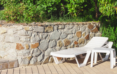 White plastic chaise lounge and table on a tiled surface next to a stone wall under green trees on a bright sunny day