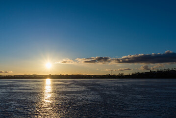 Winter landscape with frozen lake at sunrise or sunset. Lake glistening ice reflect a sun.Forest in the background.