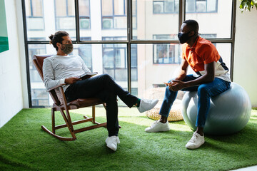 Young diverse male colleagues in masks communicating in recreation area