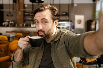 White brunette man drinking coffee while taking selfie photo in cafe