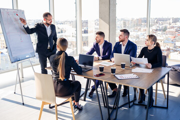 Confident businessman makes a presentation of a new project in the boardroom at a company meeting. Beautiful auditors talk with different partners about the business using a whiteboard and graphs.