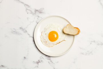Huevo frito sobre un plato redondo blanco junto a un trozo de pan sobre una mesa de mármol. Vista superior. Copy spce