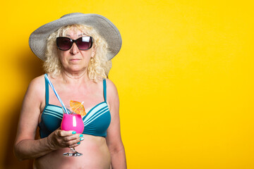 Old woman in swimsuit wearing hat and sunglasses holding cocktail on yellow background.