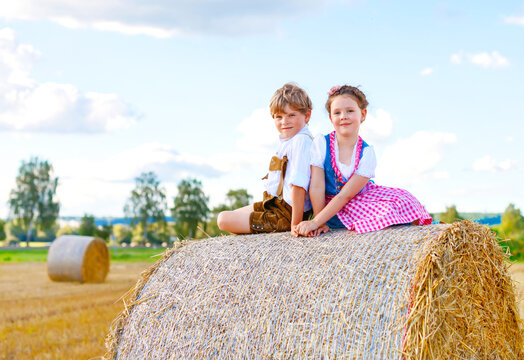 Two kids in traditional Bavarian costumes in wheat field. German children sitting on hay bale during Oktoberfest. Boy and girl play at hay bales during summer harvest time in Germany. Best friends.