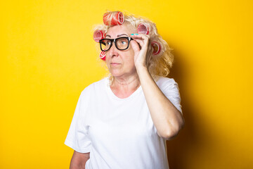 Pensive old woman with curlers in glasses looks to the side on a yellow background