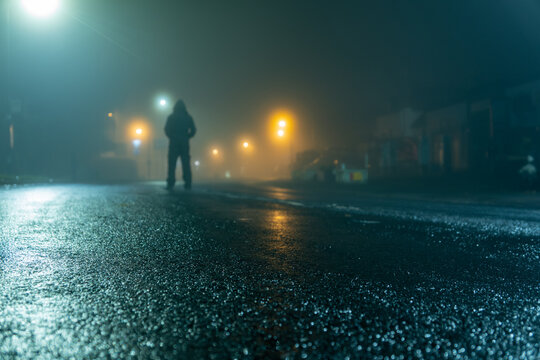 A low angle of a hooded figure standing in a street at night