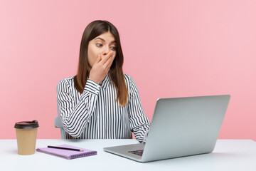 Shocked woman office worker covering mouth communicating on video call, looking at laptop screen with terrified expression, found scary secret. Indoor studio shot isolated on pink background