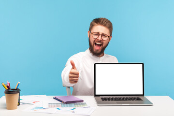 Extremely happy satisfied man office manager showing thumbs up sitting at workplace with empty screen laptop, satisfied with new pc, advertising area. Indoor studio shot isolated on blue background