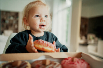beautiful little baby girl with blonde blue eyes. eats delicious fresh donuts of different flavors and colors. Happy childhood, good mood. She is dressed in a comfortable children's jacket.