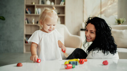The child plays with children's wooden colored toys for development. Portrait of cute little blonde girl and mom. A cheerful mood. A happy family spends time at home. Children learn at home.
