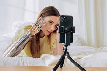 Blonde young woman shows contemporary bionic arm to camera shooting video for blog with phone on...