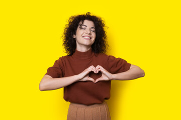 Pleased curly haired woman is gesturing heart with hands posing on a yellow studio wall