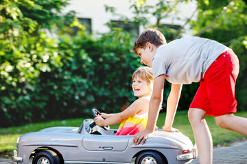 Two happy children playing with big old toy car in summer garden, outdoors. Kid boy pushing and driving car with little toddler girl, cute sister inside. Laughing and smiling kids. Lovely family