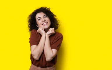Lovely curly haired woman is smiling at camera gesturing pleasure on a yellow studio wall