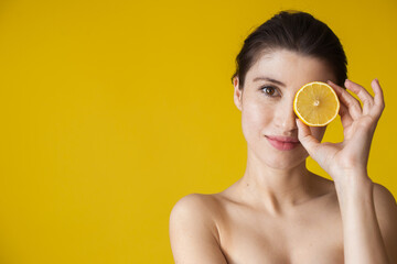 Brunette woman with freckles and bare shoulders is covering her eye with a lemon posing on a yellow studio wall with free space