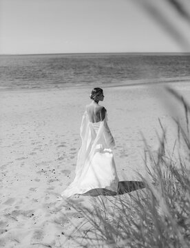 A blonde haired woman standing with her back position on the beach wearing a long bridal gown
