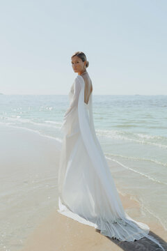 A blonde haired woman standing with her back position on the beach wearing a long bridal gown