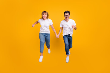 Fototapeta premium Full length photo of funny sweet two persons dressed white t-shirt running jumping holding arms isolated yellow color background