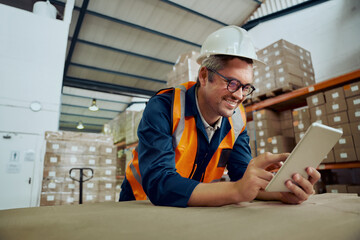 Smiling factory worker leaning on table at warehouse looking at digital tablet