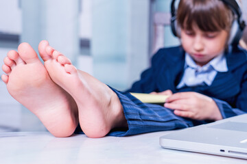 Humorous portrait of cute little business child girl with bare feet works remotely with laptop....