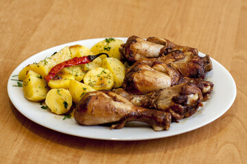 Baked chicken drumsticks with boiled potatoes, which are sprinkled with dill and decorated with pickled hot pepper. Everything is served on a white plate that lies on a light brown table.