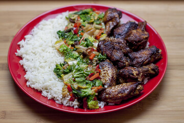 A Chinese-style dish based on baked wings served with rice and vegetables in a thick and aromatic sauce. Everything served on a red plate lying on a wooden table.