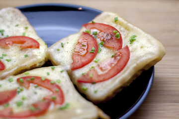 Egg-dipped toast bread and fried with melted cheese, tomato, and chives. Served on a shiny black plate lying on a wooden table.