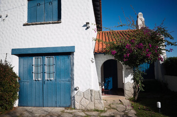 Fototapeta premium Mar del Plata, Buenos Aires, Argentina - May, 2020: Mediterranean style house with white walls, orange tiles and blue shutters. Paved courtyard and blossoming tree nearby entrance.