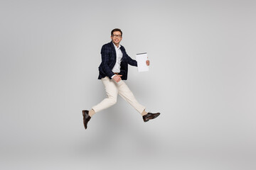 Fototapeta na wymiar full length of cheerful businessman in glasses holding paper while jumping on grey