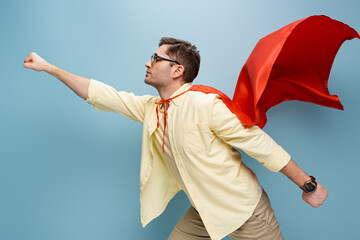side view of superhero in glasses and red cape standing with outstretched hand on blue