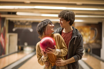 Couple standing in a bowling alley and holding a ball together. Looking each other.