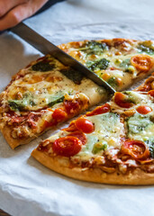 Hand cutting colorful fresh pizza hot from the oven with mozzarella cheese, pesto sauce and cherry tomatoes with a knife. Baked, delicious, gluten free pizza cut into quarters and ready to be served. 