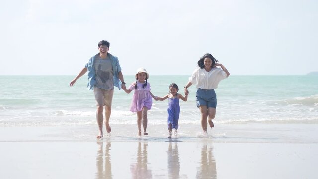 4K Happy Asian family on summer holiday vacation. Parents with two child girl kid holding hands and walking together on tropical island beach. Family enjoy and having fun outdoor lifestyle in summer.