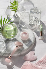 Gua Sha massager and pink quarz face roller on marble stone background. Face care products, quarz crystal ball and exotic palm leaves. Towel, skincare tonic, fragrance, soap, essential oil and serum.