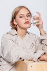 Smiling Young Woman with glass of Water, beautiful girl in beige raincoat posing in Studio