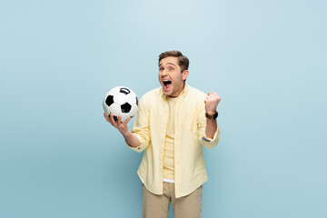 young amazed man in yellow shirt holding football and cheering on blue